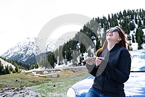 young lady sitting on a bonnet of a car using smart phone in the mountains. Happy Girl sending message with sun glasses