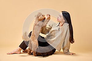 Young lady sitting on floor and feeds her purebred playful and cute poodle dog against sandy color studio background.