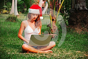 Young lady is sitting on the grass with a laptop in a christmas hat