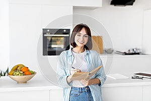 Young lady smiling and taking notes, holding notepad and pen, standing in kitchen interior, planning her day