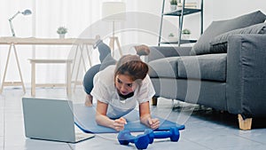 Young lady in sportswear exercises doing working out planks with a leg outstretched and using laptop to watching yoga video
