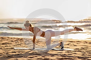 Young lady in sportswear practicing yoga by sea, doing plank exercise, enjoying workout outdoors at sunset, side view