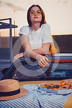 Young lady in sunglasses, white t-shirt is posing in yellow car cabriolet with pizza, striped cloth and hat on its trunk