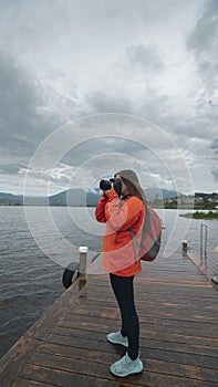 Young Latin American woman dressed in orange jacket with backpack seen from the front taking photos on a wooden pier on a lake