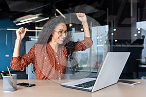 A young Latin American woman freelancer sits in the office with a laptop, raised her hands, rejoices