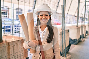 Young latin architect girl smiling happy holding blueprints standing at the city