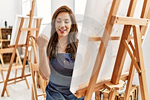 Young latin artist woman painting at art studio pointing thumb up to the side smiling happy with open mouth