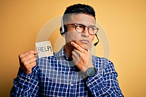 Young latin call center agent man overworked holding help message paper using headset serious face thinking about question, very