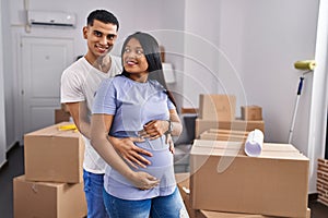 Young latin couple expecting baby hugging each other standing at new home