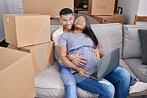 Young latin couple expecting baby touching belly using laptop at new home