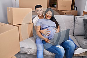 Young latin couple expecting baby touching belly using laptop at new home