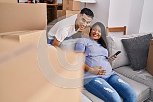Young latin couple expecting baby touching belly using smartphone at new home
