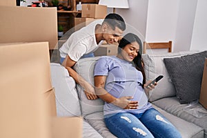Young latin couple expecting baby touching belly using smartphone at new home