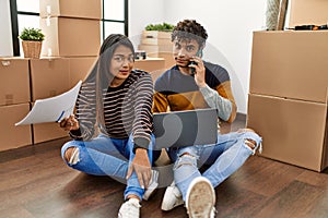 Young latin couple using laptop and talking on the smartphone sitting on the floor at new home