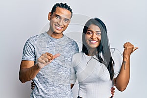 Young latin couple wearing casual clothes pointing to the back behind with hand and thumbs up, smiling confident