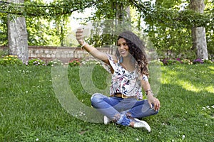 Young latin girl with dark skin and long curly hair taking a selfie with her smartphone sitting in a park