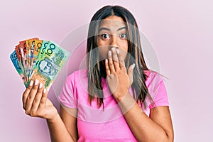 Young latin girl holding australian dollars covering mouth with hand, shocked and afraid for mistake