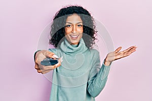 Young latin girl holding television remote control celebrating achievement with happy smile and winner expression with raised hand