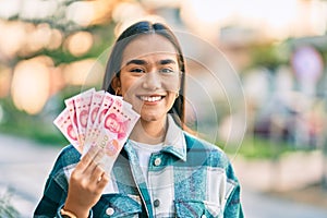 Young latin girl smiling happy holding chinese yuan banknotes at the city