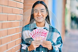 Young latin girl smiling happy holding chinese yuan banknotes at the city