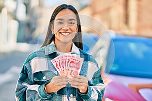 Young latin girl smiling happy holding chinese yuan banknotes at the city