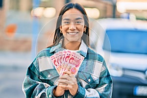 Young latin girl smiling happy holding chinese yuan banknotes at the city
