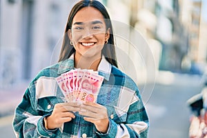 Young latin girl smiling happy holding chinese yuan banknotes at the city