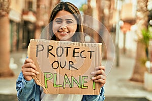Young latin girl smiling happy holding protect our planet banner at the city