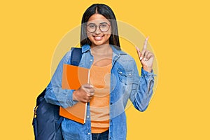 Young latin girl wearing student backpack and holding books with a big smile on face, pointing with hand finger to the side