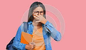 Young latin girl wearing student backpack and holding books smelling something stinky and disgusting, intolerable smell, holding