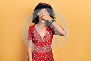 Young latin girl wearing summer dress covering eyes with hand, looking serious and sad