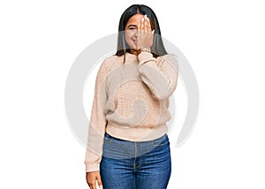 Young latin girl wearing wool winter sweater covering one eye with hand, confident smile on face and surprise emotion