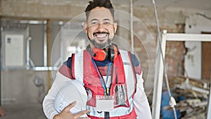 Young latin man builder smiling confident holding hardhat at construction site