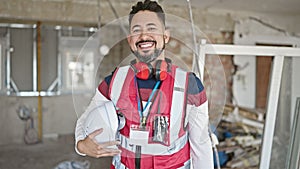 Young latin man builder smiling confident holding hardhat at construction site