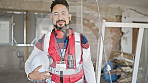 Young latin man builder smiling confident holding hardhat at construction site