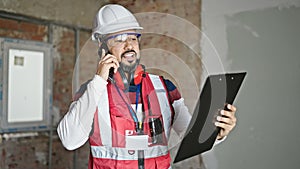 Young latin man builder talking on smartphone reading document at construction site