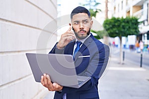 Young latin man business worker using laptop talking on smartphone at street