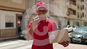 Young latin man delivery worker holding package doig money gesture with finger at street