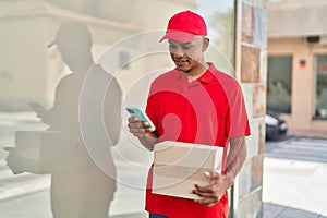 Young latin man delivery worker holding package using smartphone at street