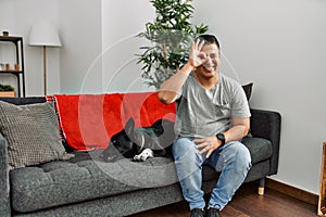 Young latin man and dog sitting on the sofa at home doing ok gesture with hand smiling, eye looking through fingers with happy