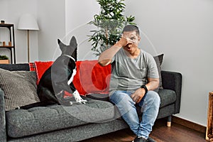 Young latin man and dog sitting on the sofa at home peeking in shock covering face and eyes with hand, looking through fingers