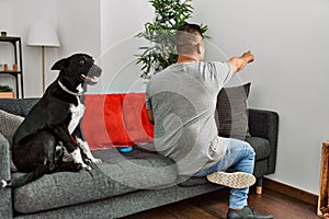 Young latin man and dog sitting on the sofa at home posing backwards pointing ahead with finger hand