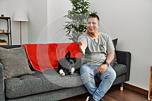 Young latin man and dog sitting on the sofa at home smiling friendly offering handshake as greeting and welcoming