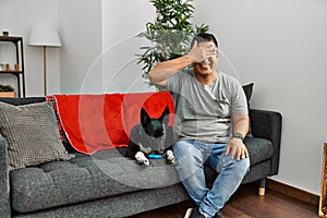 Young latin man and dog sitting on the sofa at home smiling and laughing with hand on face covering eyes for surprise