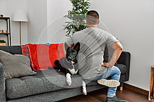 Young latin man and dog sitting on the sofa at home standing backwards looking away with arms on body