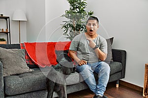 Young latin man and dog sitting on the sofa at home surprised pointing with finger to the side, open mouth amazed expression