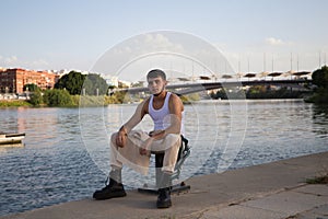 Young latin man dressed in beige pants, white t-shirt and black boots sitting on the mooring at the river pier in seville. The man