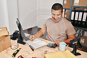 Young latin man ecommerce business worker using calculator writing on notebook at office