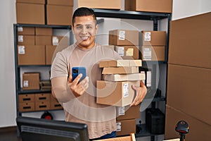 Young latin man ecommerce business worker using smartphone holding packages at office