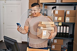 Young latin man ecommerce business worker using smartphone holding packages at office